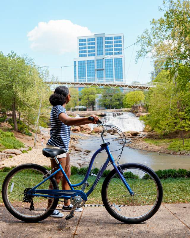 Woman with medium skin tone, blue and white striped shirt, and white shorts standing with a bike admiring the Liberty Bridge at Falls Park on the Reedy in Greenville, SC on a sunny summer day.
