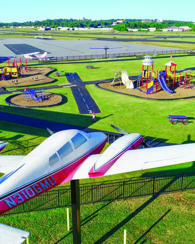 A red and white airplane sculpture stands in the foreground of a playground with a wide airport runway in the background.