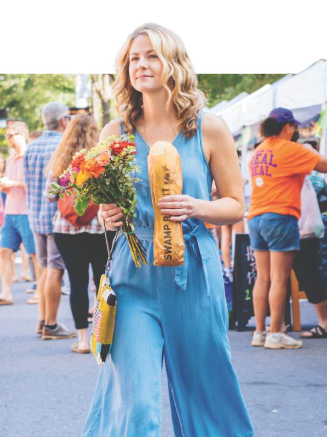 Fair-skinned woman in a denim jumpsuit holding a loaf of Swamp Rabbit Grocery and Cafe stecca bread and a bouquet of wildflowers while shopping at the TD Saturday Market in Downtown Greenville, SC on a sunny Saturday morning with other shoppers and vendor tents in the background.