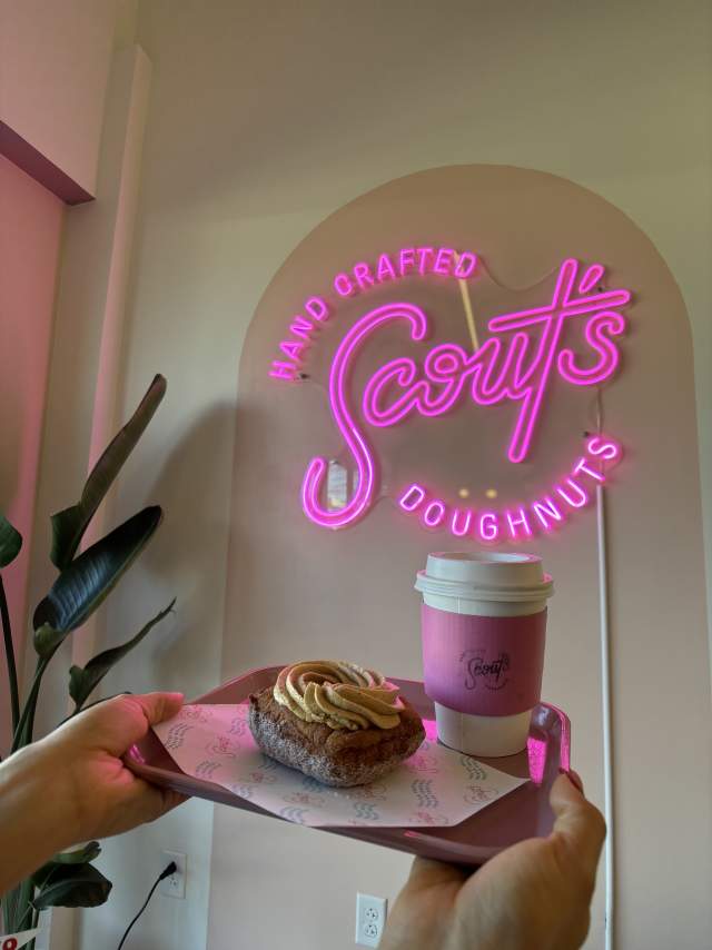Person's hands holding a tray with a fluffy donut with frosting on top and a coffee cup with a pink sleeve and the Scout's Donuts logo in front of a pink neon light up sign of the Scout's Donuts logo.