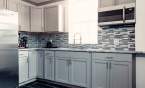 White and grey kitchen at Kings Collective with a silver fridge and microwave, grey lower cabinets, white upper cabinets and a grey/white/black tile backsplash and black tile floor.