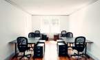 Six black desks and black office chairs in a bright white coworking office space with hardwood floors and several windows.