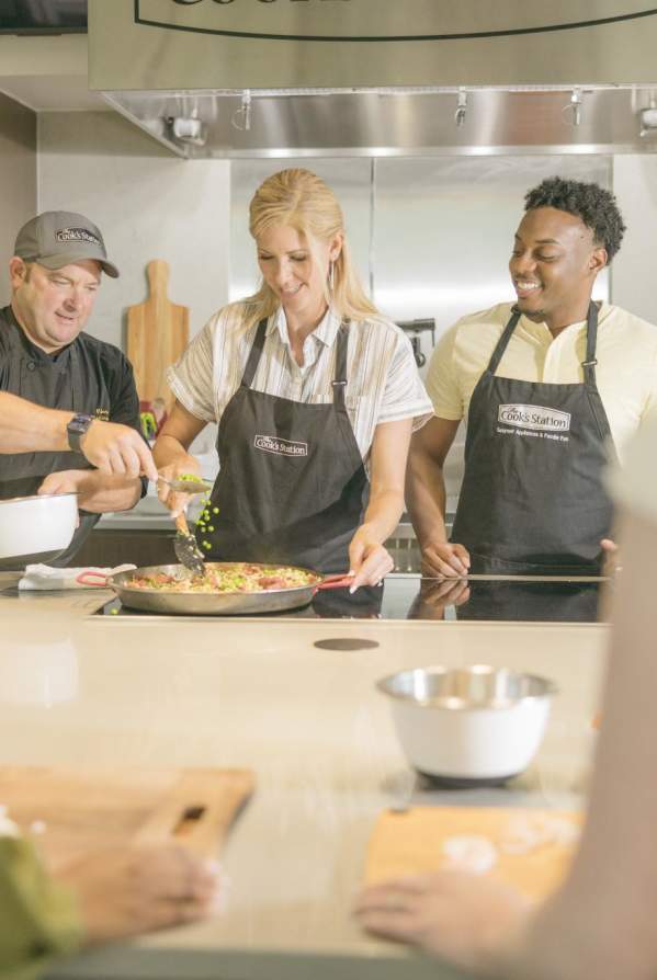 Group of men and women at a cooking class with a chef demonstrating how to prepare a dish on a stove.
