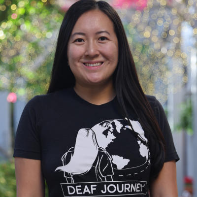 A woman in a black t-shirt with a "DEAF JOURNEY" logo smiles at the camera, with a blurred background of greenery and lights.