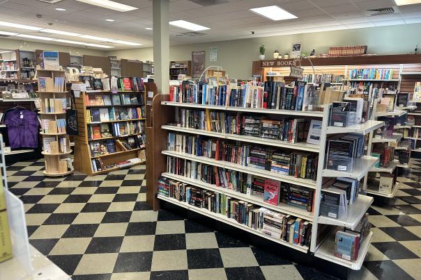 Interior image of As the Page Turns bookstore in Travelers Rest with many shelves of fiction books pictured in a shop with a black and white checkerboard floor and overhead lighting.