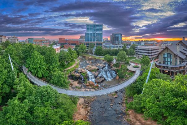 An aerial view of downtown Greenville, featuring the iconic Liberty Bridge spanning over Falls Park, with lush greenery and urban buildings in the background.
