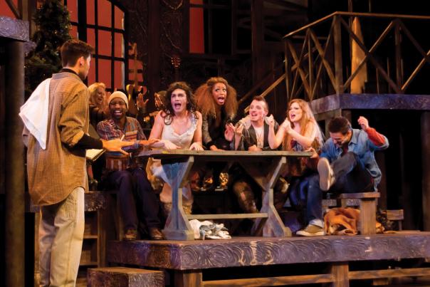 A lively scene from 'Rent,' with actors in 80s costumes gathered around a central table, singing passionately