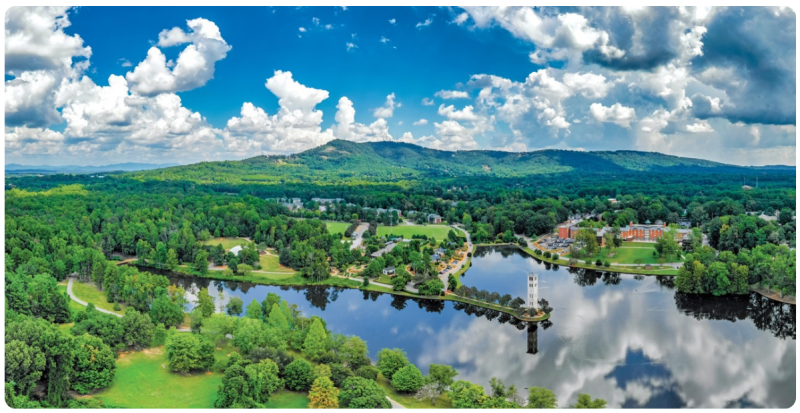 Lush green parkland surrounds a tranquil lake with a prominent tower, all set against a backdrop of rolling hills under a cloudy blue sky.