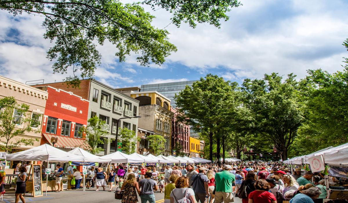 Downtown Greenville farmers market with many vendors set up and a large crowd of shoppers walking by the stands.