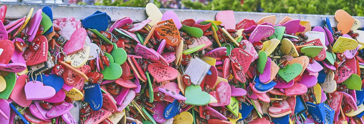 A dense collection of colorful heart-shaped locks adorns a fence, with blurred green foliage visible in the background.