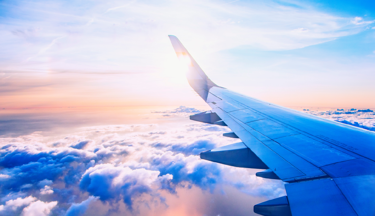 An airplane wing extends into a sky filled with soft clouds and a glowing sun.