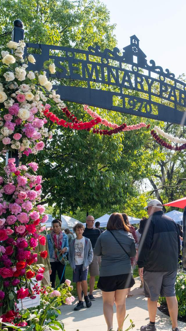 Visitors walk beneath the flower-covered Seminary Park archway at the Indiana Peony Festival. The arch is decorated with pink, white, and red peonies, and crowds explore vendor tents in the sunny park beyond.