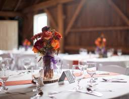 A close-up of a rustic wooden table with a purple floral centerpiece in a glass jar, set with white plates and glasses in a barn-like setting.
