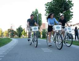 Three people ride white bike-share bicycles along a paved path, smiling as they pedal side by side on a sunny day.