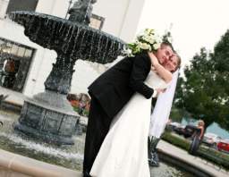 A smiling man and woman in wedding attire embracing in front of a stone water fountain.