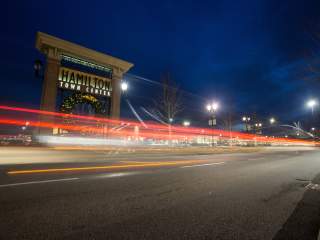Illuminated Hamilton Town Center entrance at night with holiday decor, as long-exposure car light trails streak along the curved road under a deep blue sky.