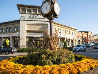 A street clock in the center of a roundabout, in front of multi-story buildings with storefronts and landscaping.