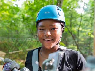 Person wearing a blue helmet and safety harness standing on an outdoor ropes course, smiling toward the camera, with trees and another participant visible in the background.