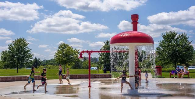 Children run and play through a splash pad at a park, dashing under a large red-and-white water feature that sprays streams downward, with trees, grass, and a partly cloudy sky in the background.