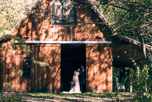 A bride standing in the doorway of a large, rustic wooden barn with the sun shining behind them.