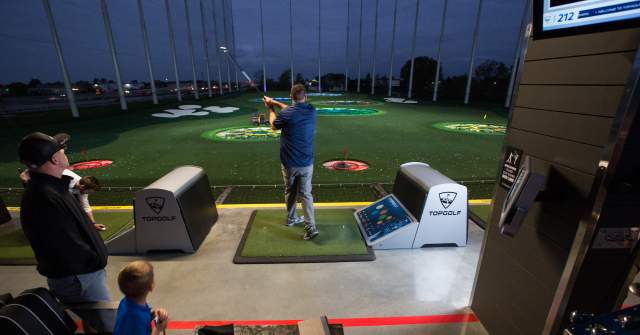 People play golf at an outdoor Topgolf bay at night, with one person swinging at the tee while others watch and a lit driving range in the background.