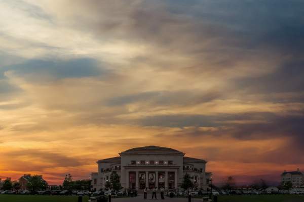 Dramatic sunset sky with layered clouds above a large, columned building silhouetted against warm orange and purple tones.