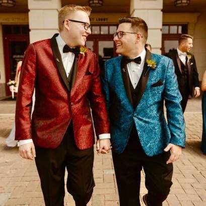 Two grooms in coordinated formal jackets—one red, one blue—holding hands and smiling at each other while walking outside, surrounded by wedding party members in formal attire.