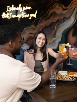 A group of four friends toasting with colorful cocktails at a table with a vibrant mural in the background, featuring food and drinks.