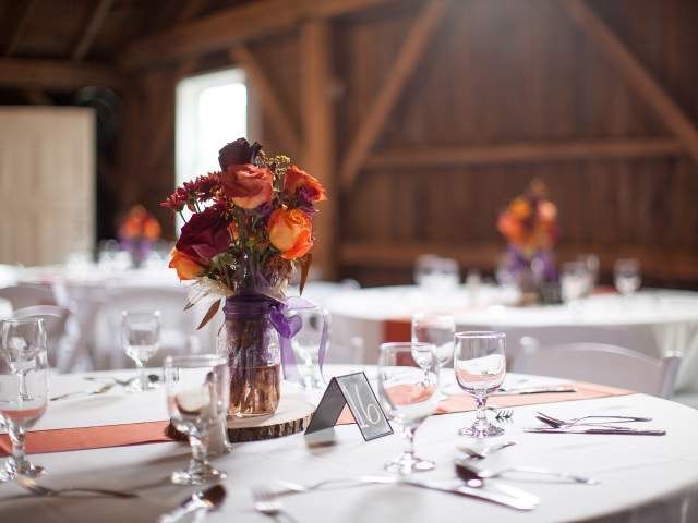 A close-up of a rustic wooden table with a purple floral centerpiece in a glass jar, set with white plates and glasses in a barn-like setting.