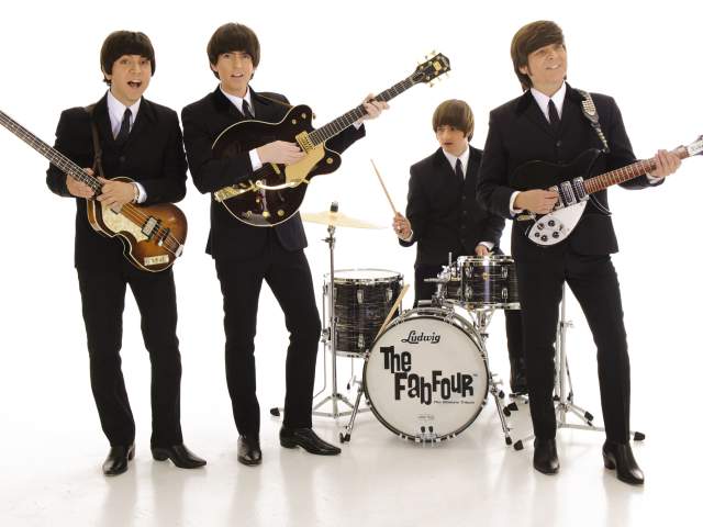 Studio photo of four musicians standing against a white background, wearing matching black suits and holding guitars and drumsticks, posed around a drum kit labeled ‘The Fab Four.’