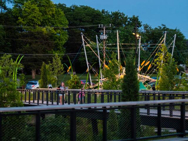 Evening view of Grand Junction Plaza during Jams at the Junction, with a bright moon in the sky above trees, string-lit structures, and people gathered around the railings and walkways.