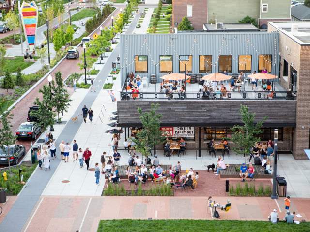A bustling outdoor dining area with patrons enjoying meals under umbrellas. Nearby, a tree-lined walkway features people strolling and socializing.