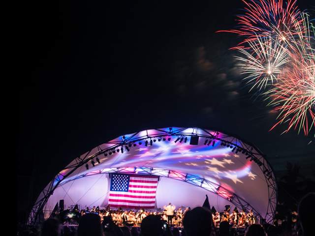 A white, shell-shaped outdoor stage lit with red, white, and blue lighting and an American flag displayed onstage. Fireworks burst above the stage against a dark night sky.