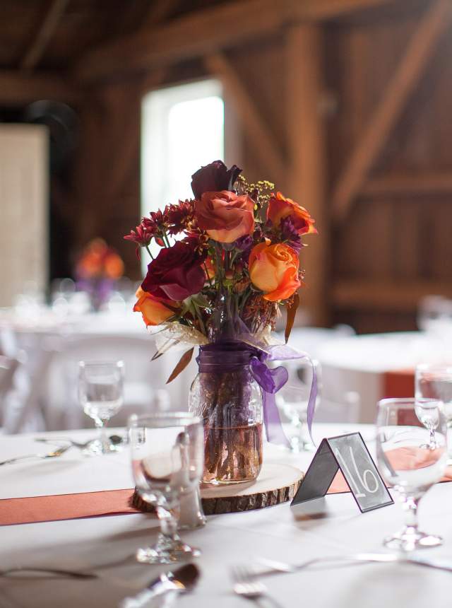 A close-up of a rustic wooden table with a purple floral centerpiece in a glass jar, set with white plates and glasses in a barn-like setting.