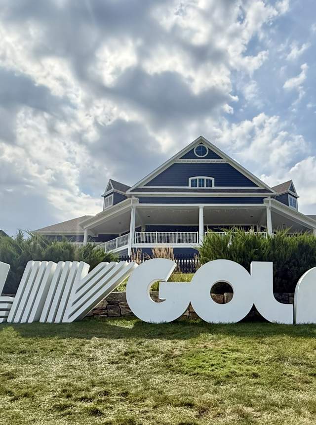 Large white “LIV Golf” letters stand on a grassy lawn in front of a clubhouse-style building under a partly cloudy sky.