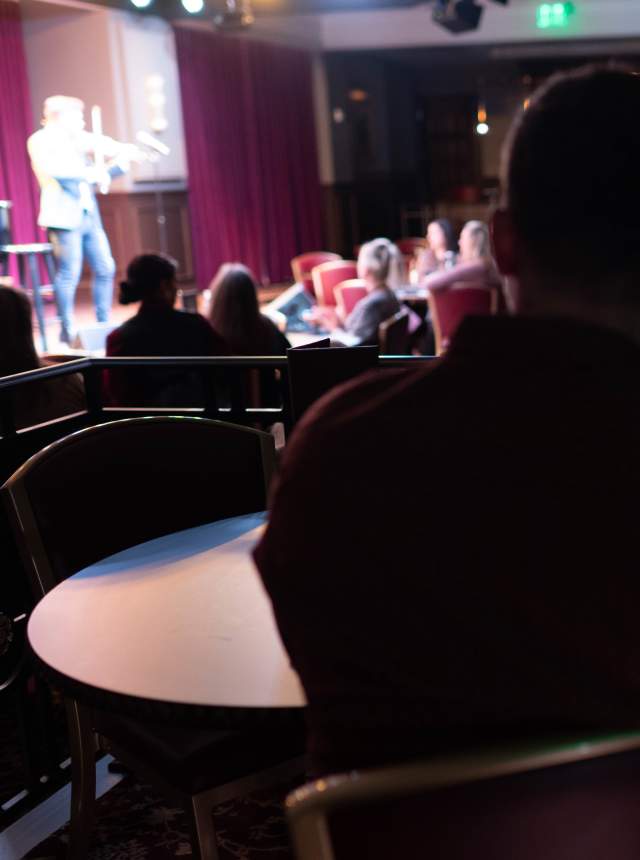 A dimly lit venue with a stage, featuring a performer in the foreground. Patrons sit at tables, enjoying the show.