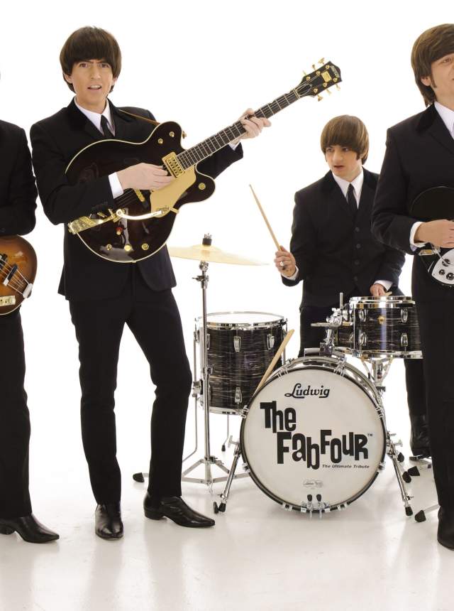 Studio photo of four musicians standing against a white background, wearing matching black suits and holding guitars and drumsticks, posed around a drum kit labeled ‘The Fab Four.’