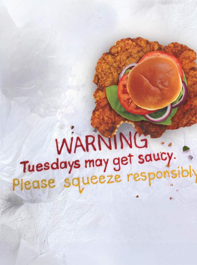 Crispy tenderloin sandwich on parchment paper with orange sauce spread underneath, paired with bold text that reads: “WARNING: Tuesdays may get saucy. Please squeeze responsibly.”