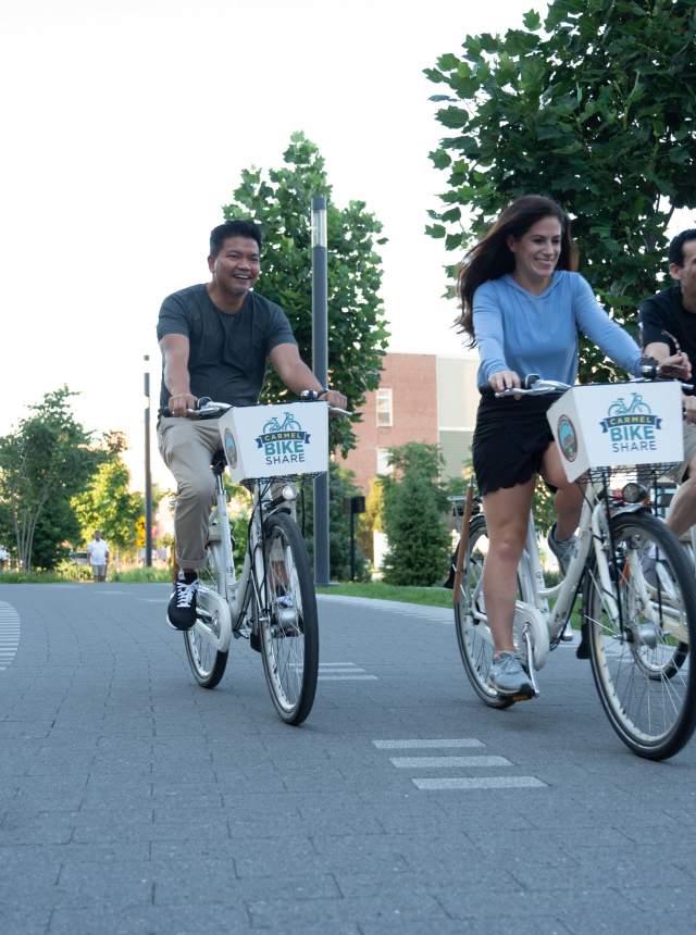 Three people ride white bike-share bicycles along a paved path, smiling as they pedal side by side on a sunny day.