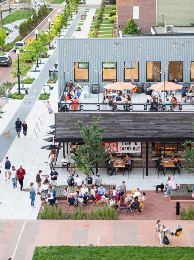 A bustling outdoor dining area with patrons enjoying meals under umbrellas. Nearby, a tree-lined walkway features people strolling and socializing.
