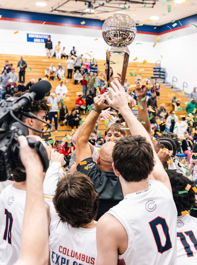 A group of high school basketball players lift a championship trophy in a gymnasium as confetti falls, surrounded by photographers and spectators in the bleachers.
