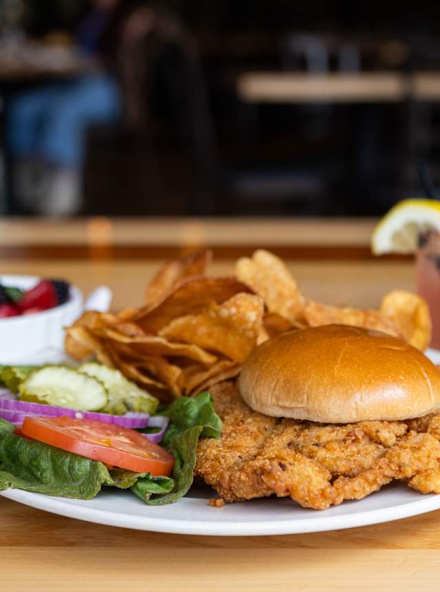 A plated breaded tenderloin sandwich with chips, fresh vegetables, a side of mixed berries, and a drink at Four Day Ray Brewing.