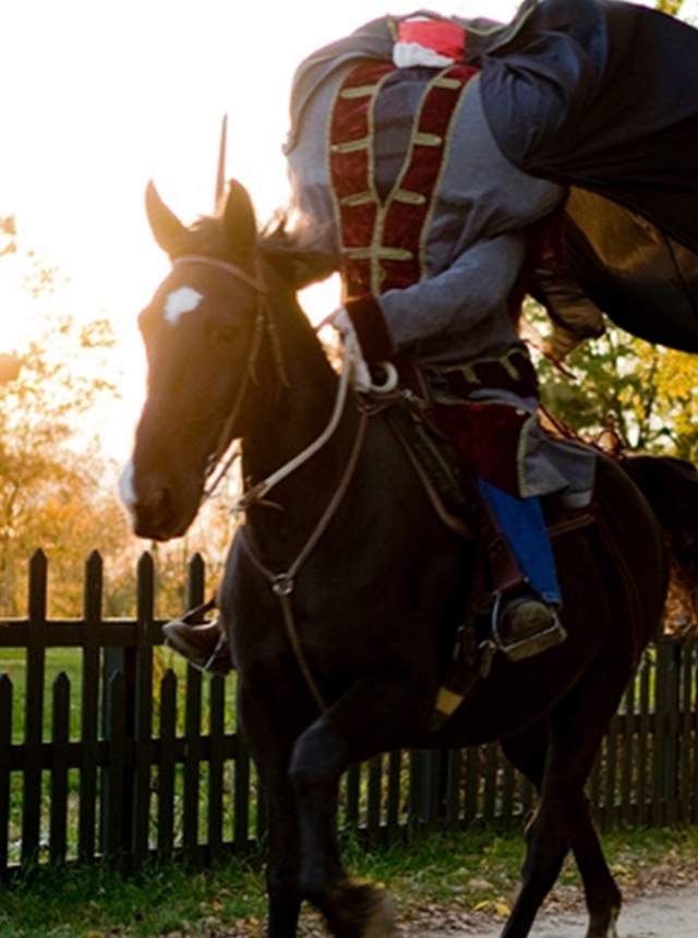 Person in period-style clothing posed as a headless rider, leaning forward on a dark horse along a dirt path beside a wooden fence. A historic-style house and trees are visible in the background, lit by warm, low-angle sunlight.