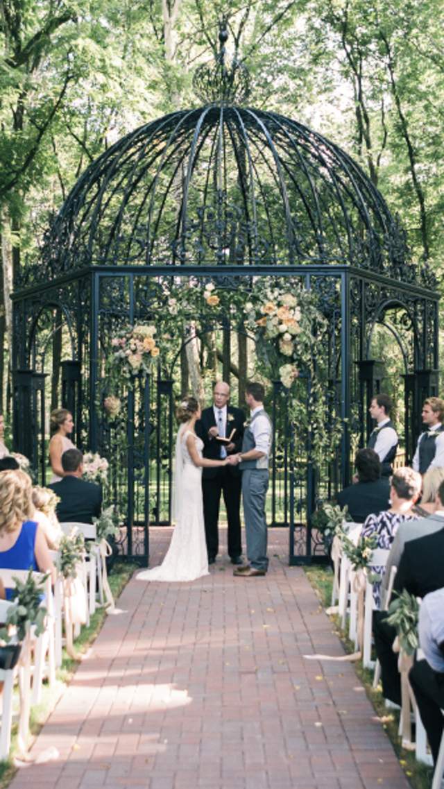 An outdoor wedding ceremony taking place under an ornate black iron gazebo surrounded by lush greenery. A large gather of people are seating facing the gazebo, with a bride and groom in the center.