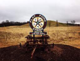 A vintage-style railroad crossing sign mounted on a wooden post with a grassy field background at Flor Fork Creek Park.