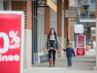 A woman carries a baby in a front-facing carrier while holding hands with a young boy. They walk past storefronts and promotional signs.