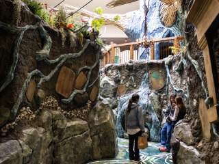 Two visitors explore a whimsical, nature-inspired indoor pathway with rocky textures, greenery, and artistic details. The scene invites adventure.