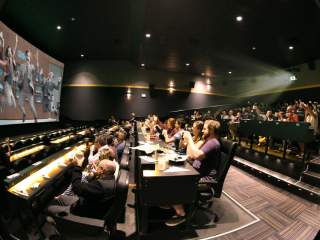 Movie theater auditorium with tiered seating and tables where guests are watching a film on a large screen while eating and drinking, with a projector beam visible and many audience members raising their hands toward the screen.