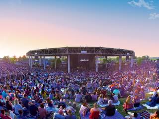 Large outdoor crowd seated on blankets and lawn chairs facing the stage at Ruoff Music Center during a sunset concert, with the amphitheater roof centered in the background.