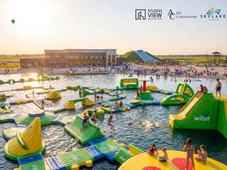 A vibrant water park scene featuring inflatable structures in a lake, with families enjoying activities under a sunset sky. A beach area is visible.